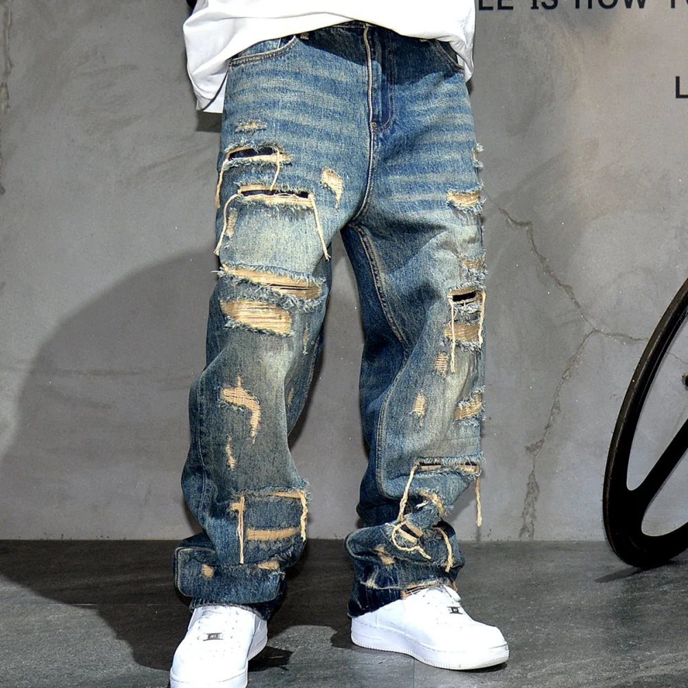 A person wearing heavily distressed baggy blue denim jeans and white sneakers against a grey wall.