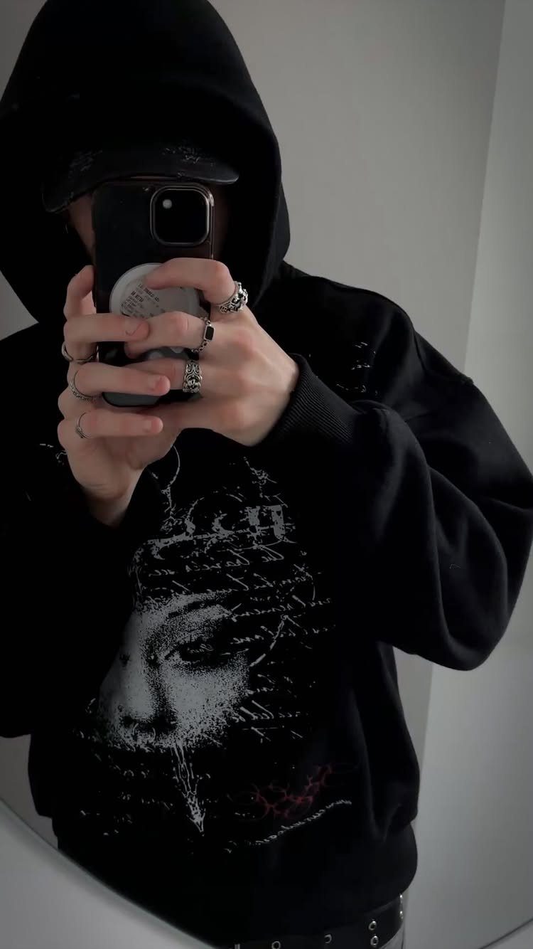 Person in a black oversized graphic hoodie taking a mirror selfie with silver rings.