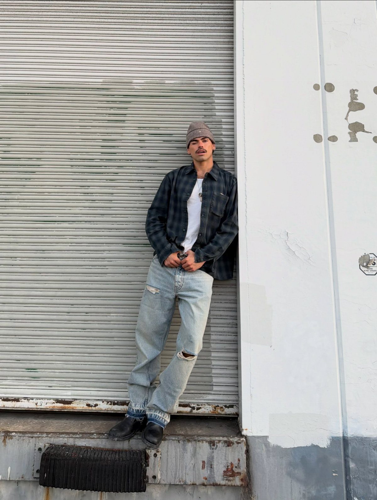 Man wearing an oversized plaid flannel shirt and baggy distressed jeans leaning against a wall.