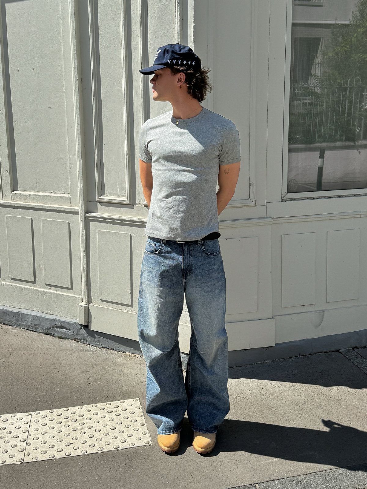 A man wearing a fitted grey t-shirt and baggy blue jeans standing against a white wall.