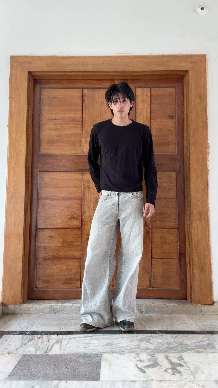 A young man posing in a black long-sleeved shirt and light grey baggy jeans.