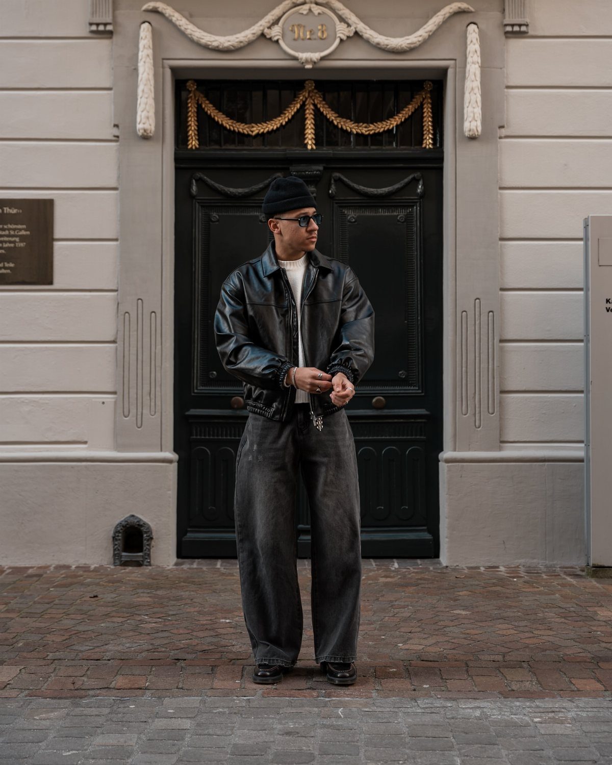 Man wearing a black leather jacket and baggy dark jeans standing before a large black door.