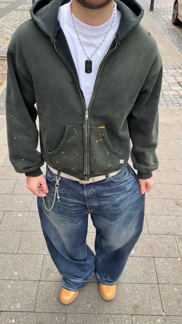 A man wearing a dark green hoodie and very baggy blue denim jeans standing outdoors.