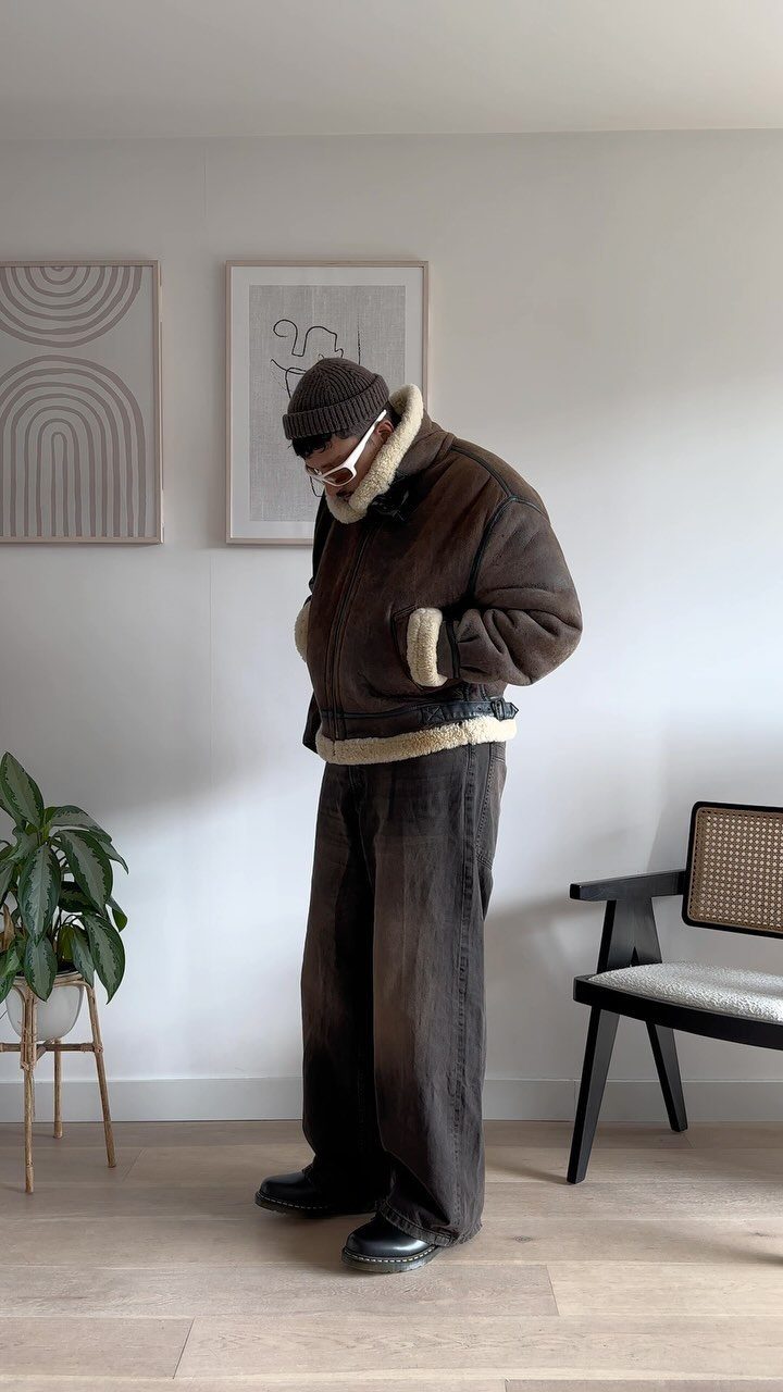 A person wearing a brown shearling jacket and baggy dark brown trousers standing in a room.