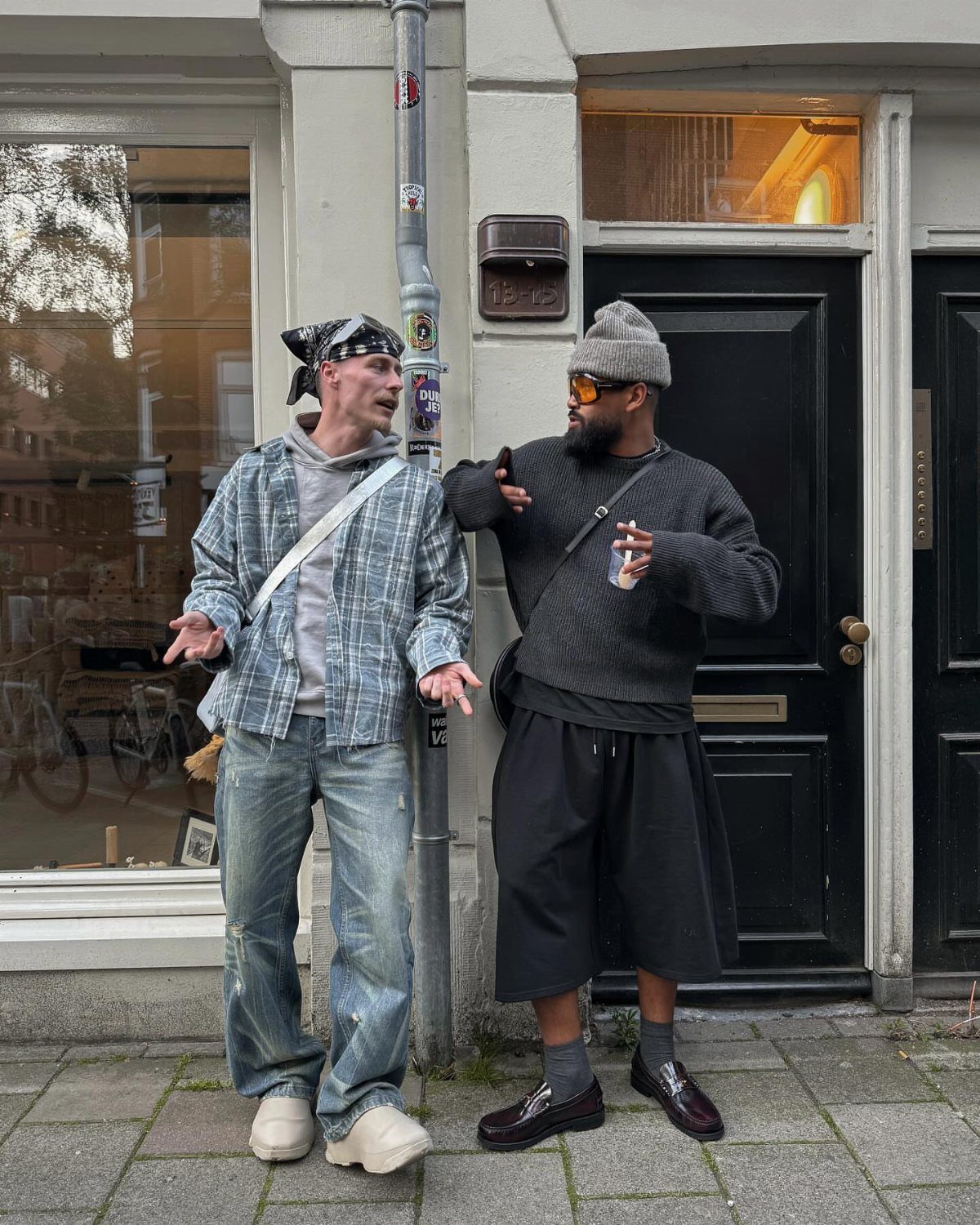 Two men in streetwear standing on a city sidewalk in front of a black door.
