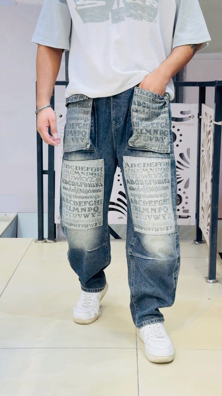 Person wearing baggy blue denim jeans with large alphabet-printed patches and white sneakers.
