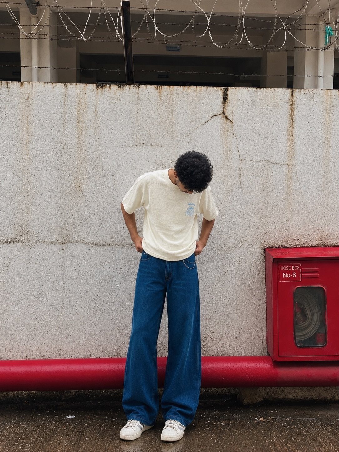 Man wearing baggy blue jeans, a cream t-shirt, and white sneakers standing against a white wall.