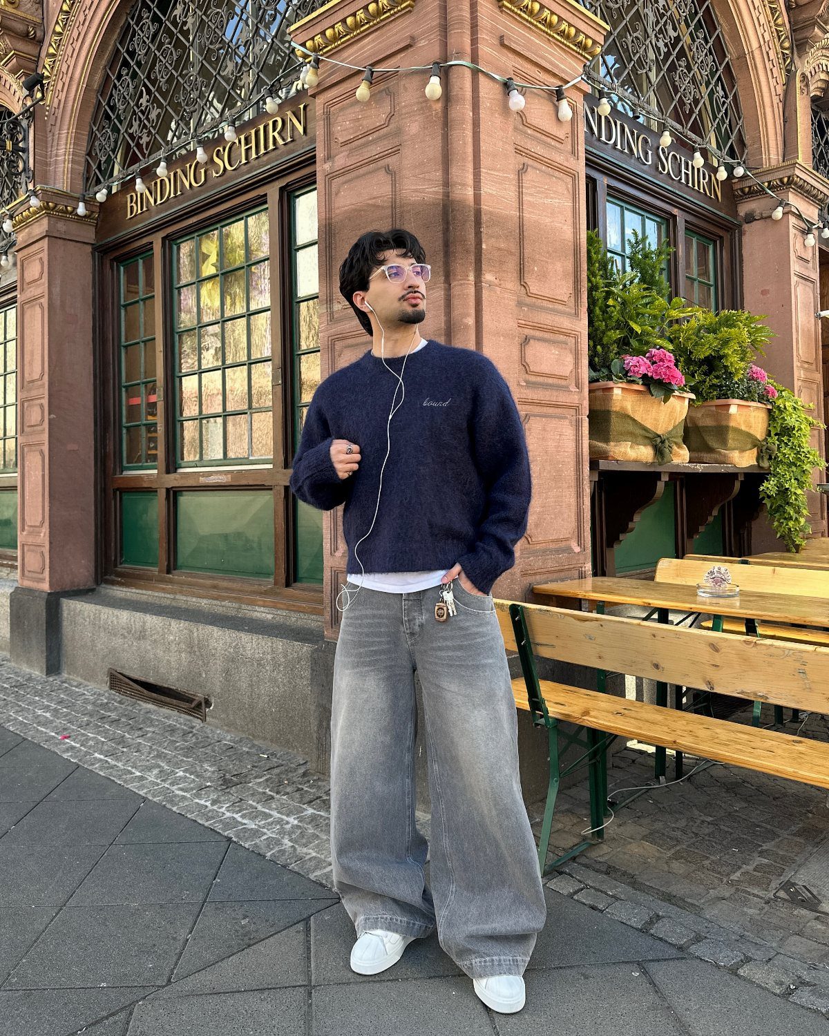 Man wearing grey baggy jeans and a dark blue sweater standing outdoors near a building.