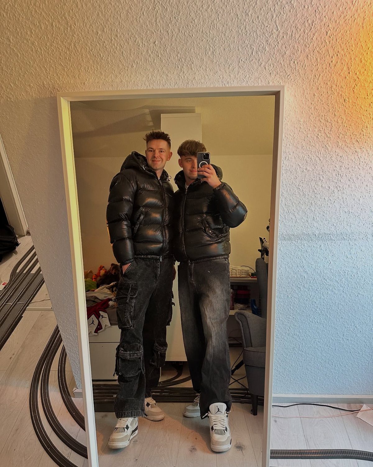 Two men wearing black puffer jackets and baggy jeans posing for a mirror selfie.