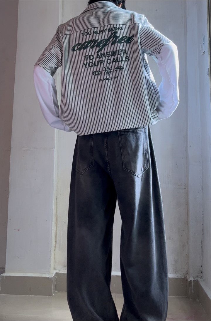 Person wearing wide baggy grey jeans and a striped shirt standing against a white wall.