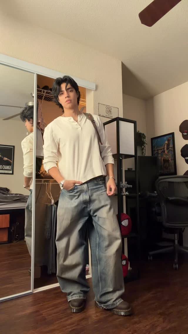 A young man posing in a room wearing a white shirt and baggy blue jeans.