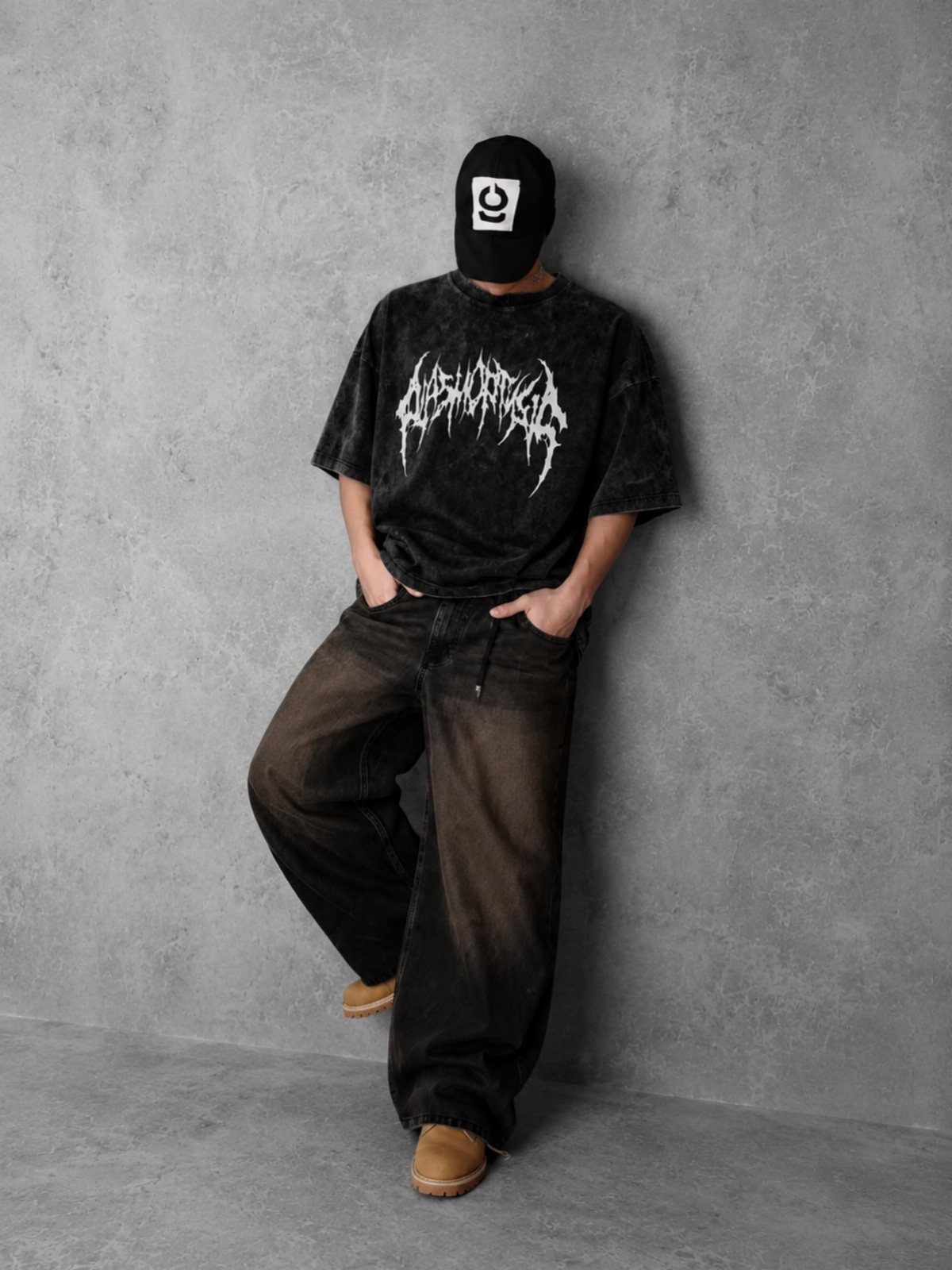 Person in a black graphic t-shirt and baggy dark jeans leaning against a grey wall.