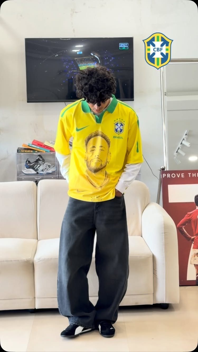 A person wearing a yellow Brazil soccer jersey and baggy black jeans standing indoors.