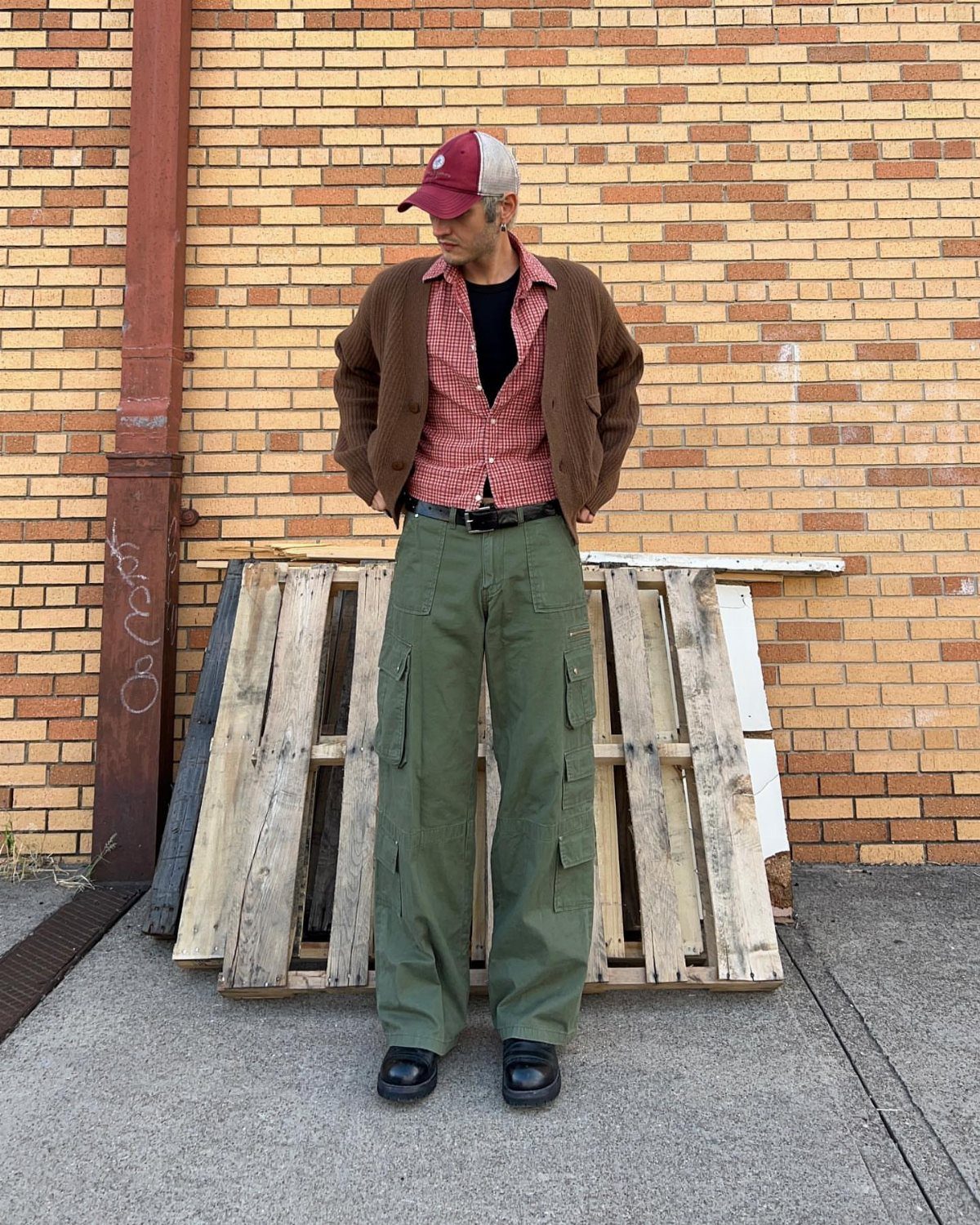 A man posing in olive green baggy cargo pants, a brown cardigan, and a red cap.
