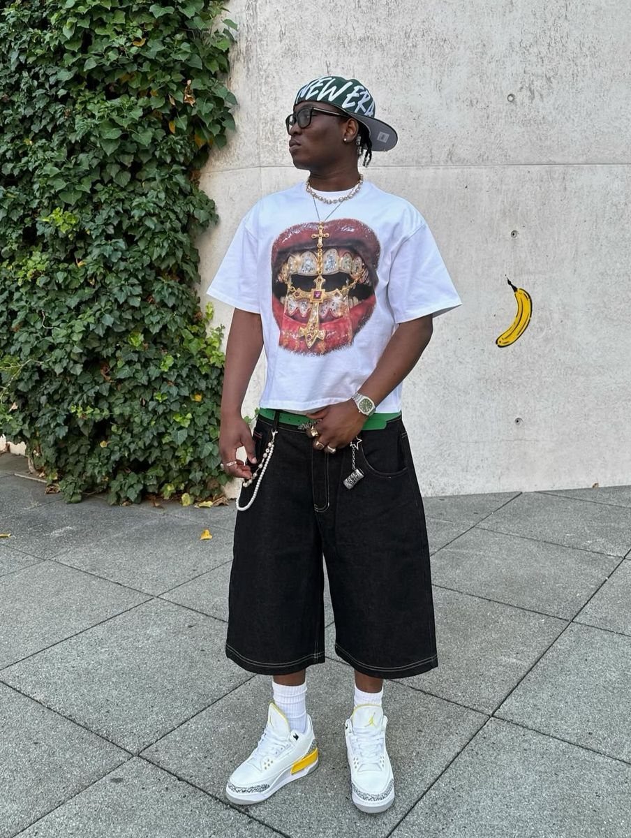 A man wearing a white graphic t-shirt and baggy black denim shorts standing outdoors.