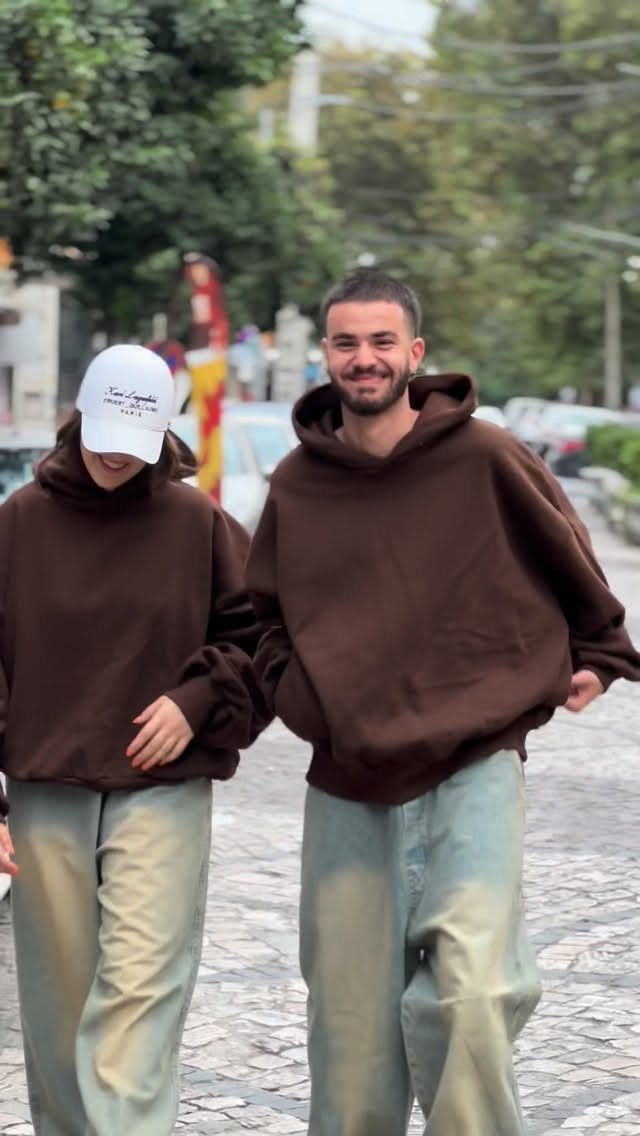 A man and woman walking in brown oversized hoodies and baggy light-colored jeans.