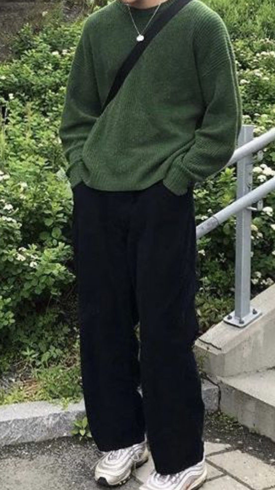 A man wearing a dark green crewneck sweater and wide-leg black trousers standing outdoors.