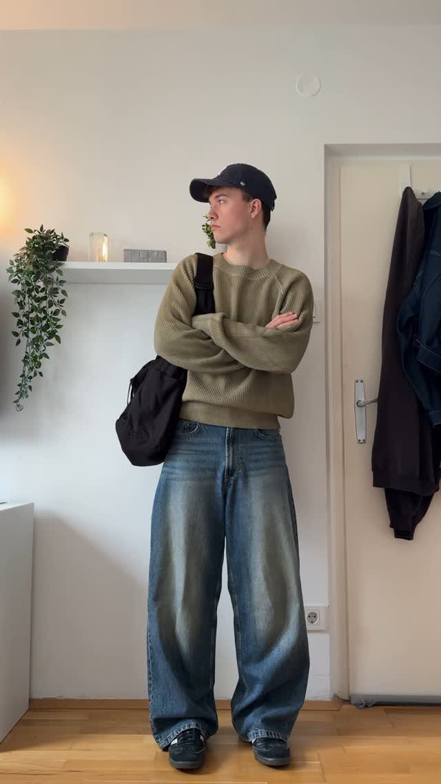 Man in olive green crewneck sweater and wide-leg blue denim jeans standing indoors.