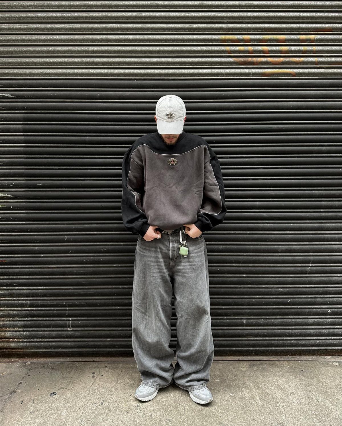 Man wearing a grey graphic sweatshirt and baggy grey jeans standing before a metal shutter.