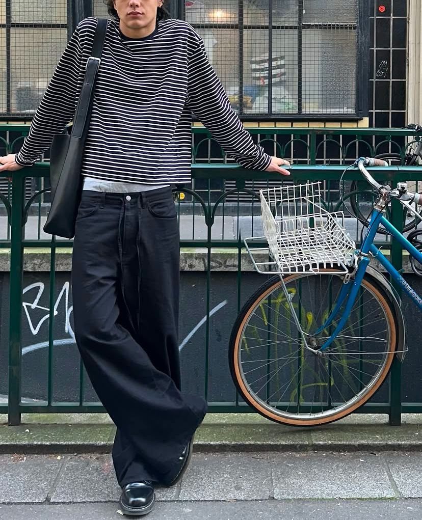 Man in a striped long-sleeve shirt and wide-leg black trousers leaning against a green fence.