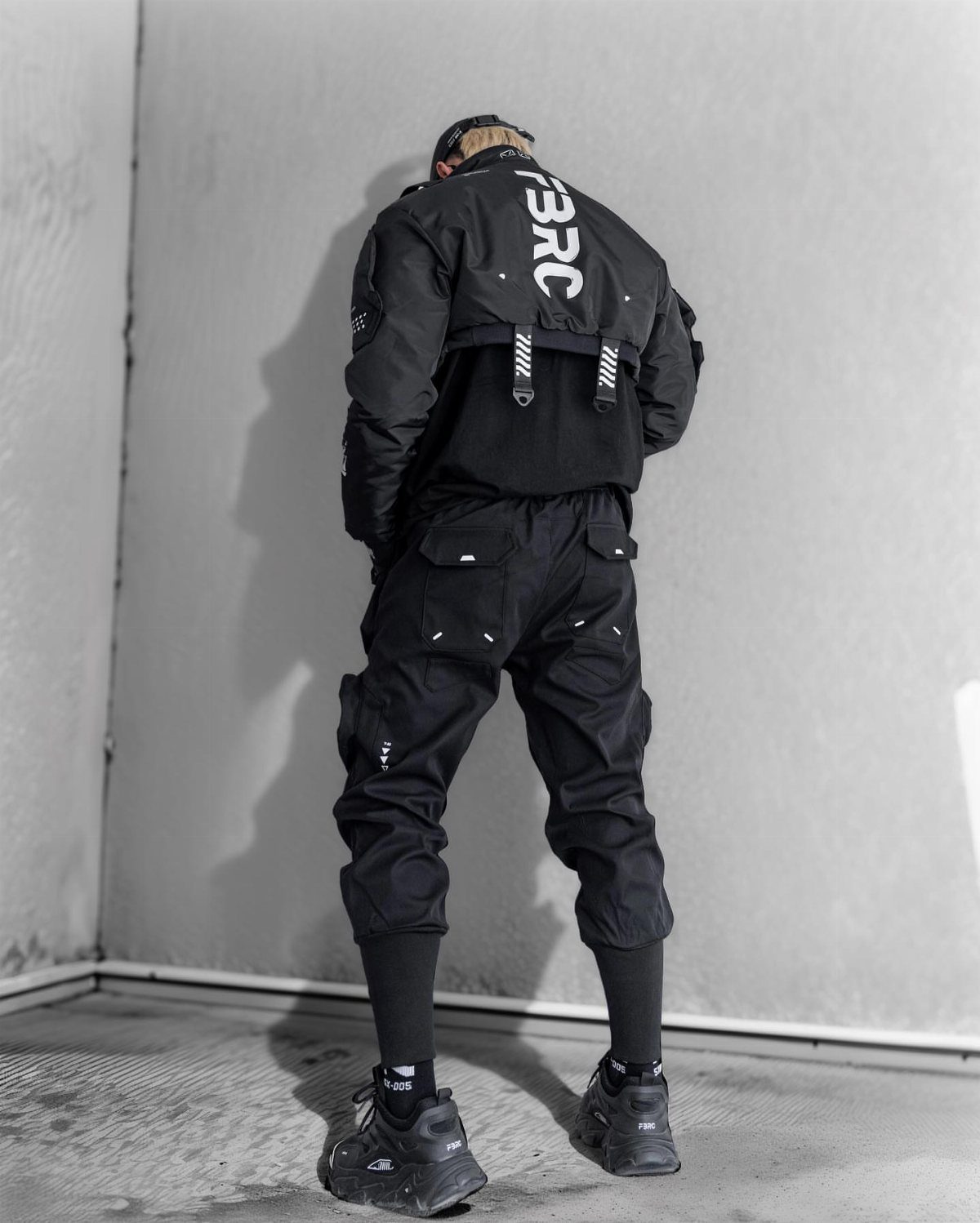 Man in black technical jacket and black cargo pants standing against a grey wall.
