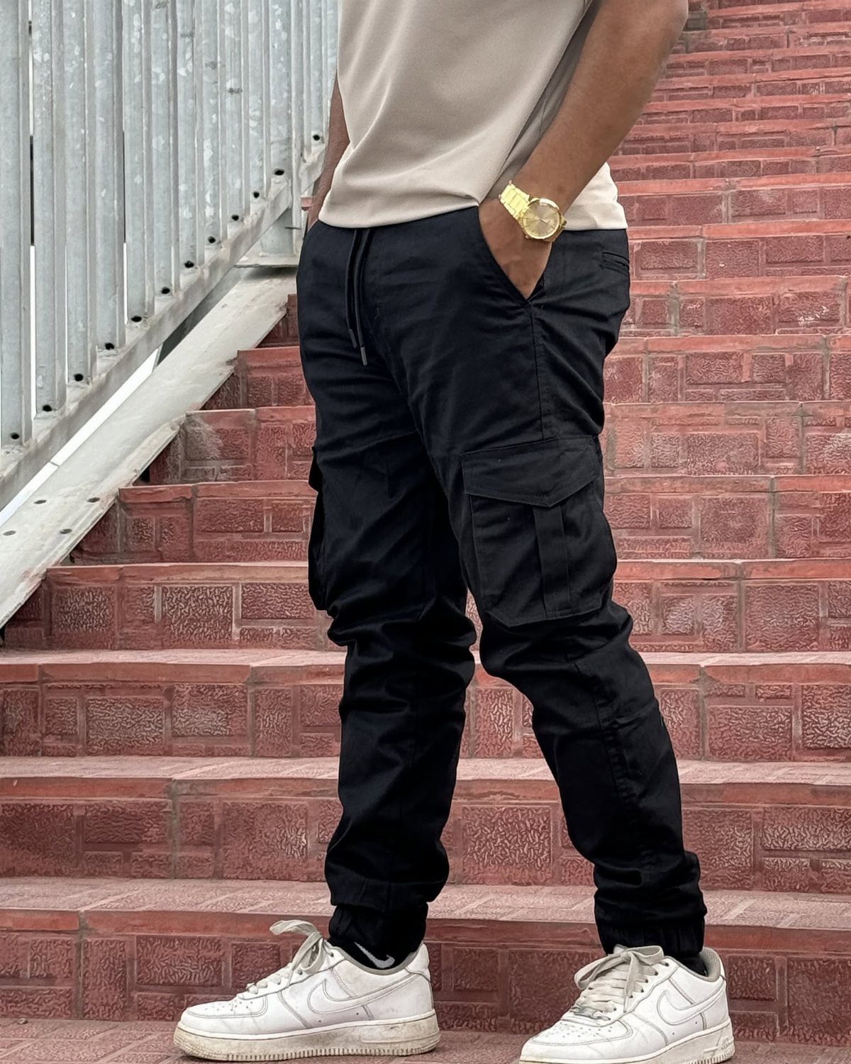 A man wearing black cargo pants, a beige t-shirt, and white sneakers standing on brick stairs.