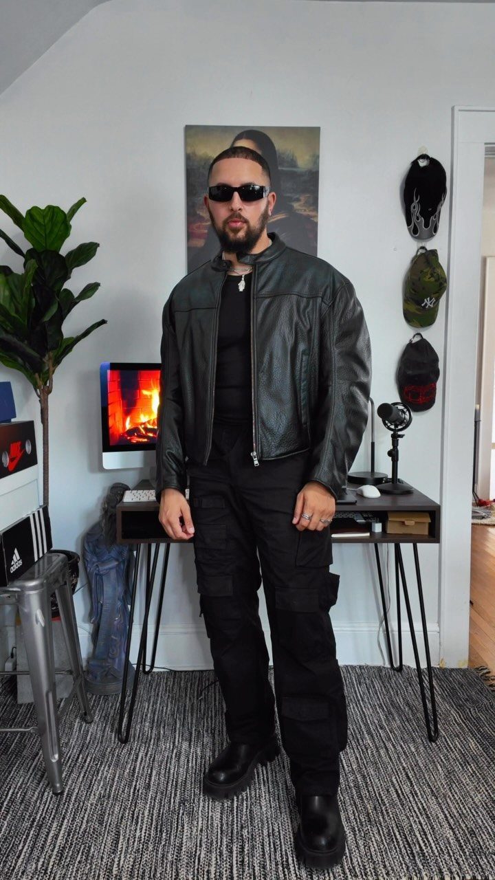 Man in black leather jacket, black cargo pants, and black Chelsea boots standing indoors.