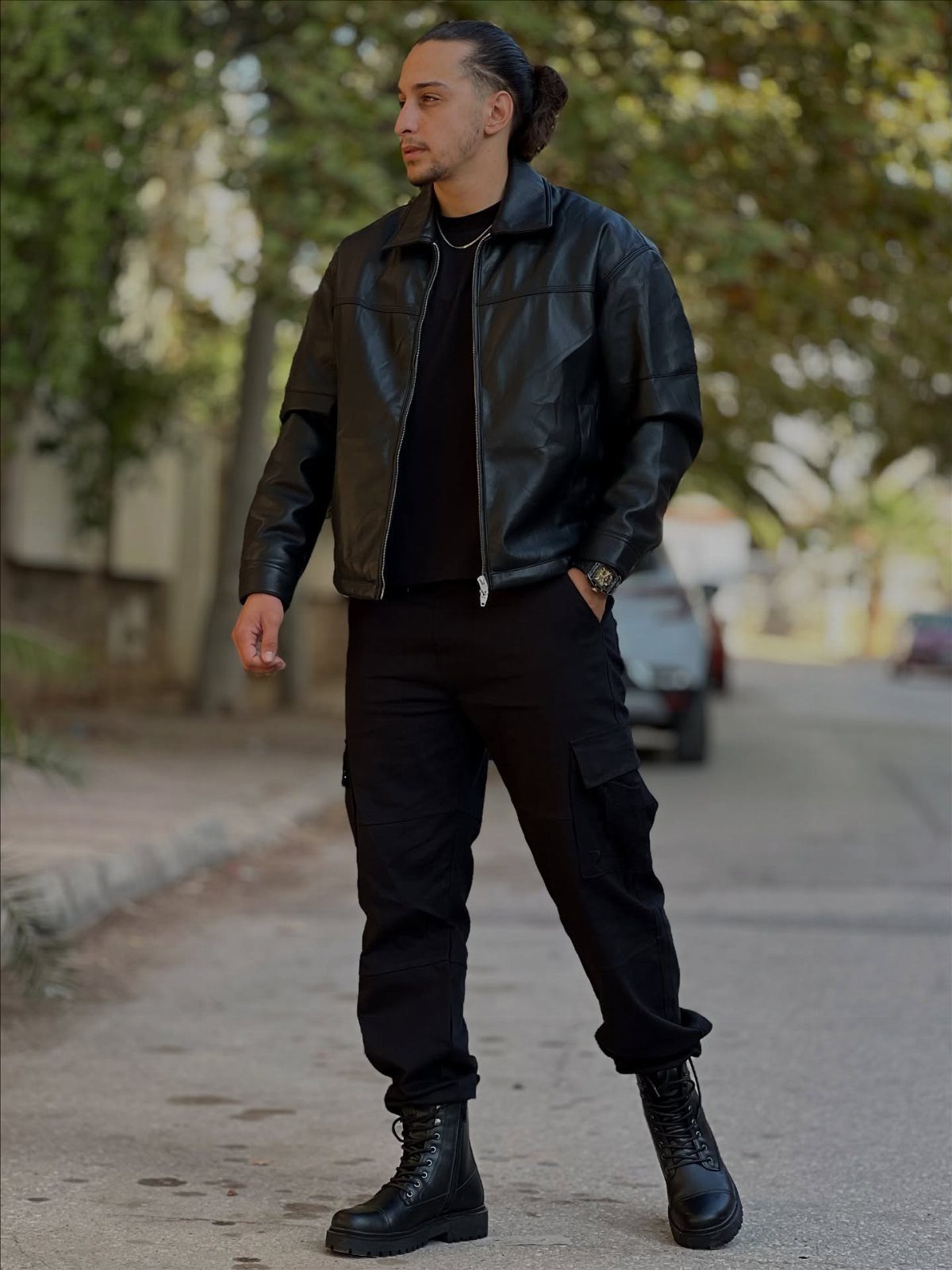 Man wearing black leather jacket, black cargo pants, and black combat boots standing on a street.