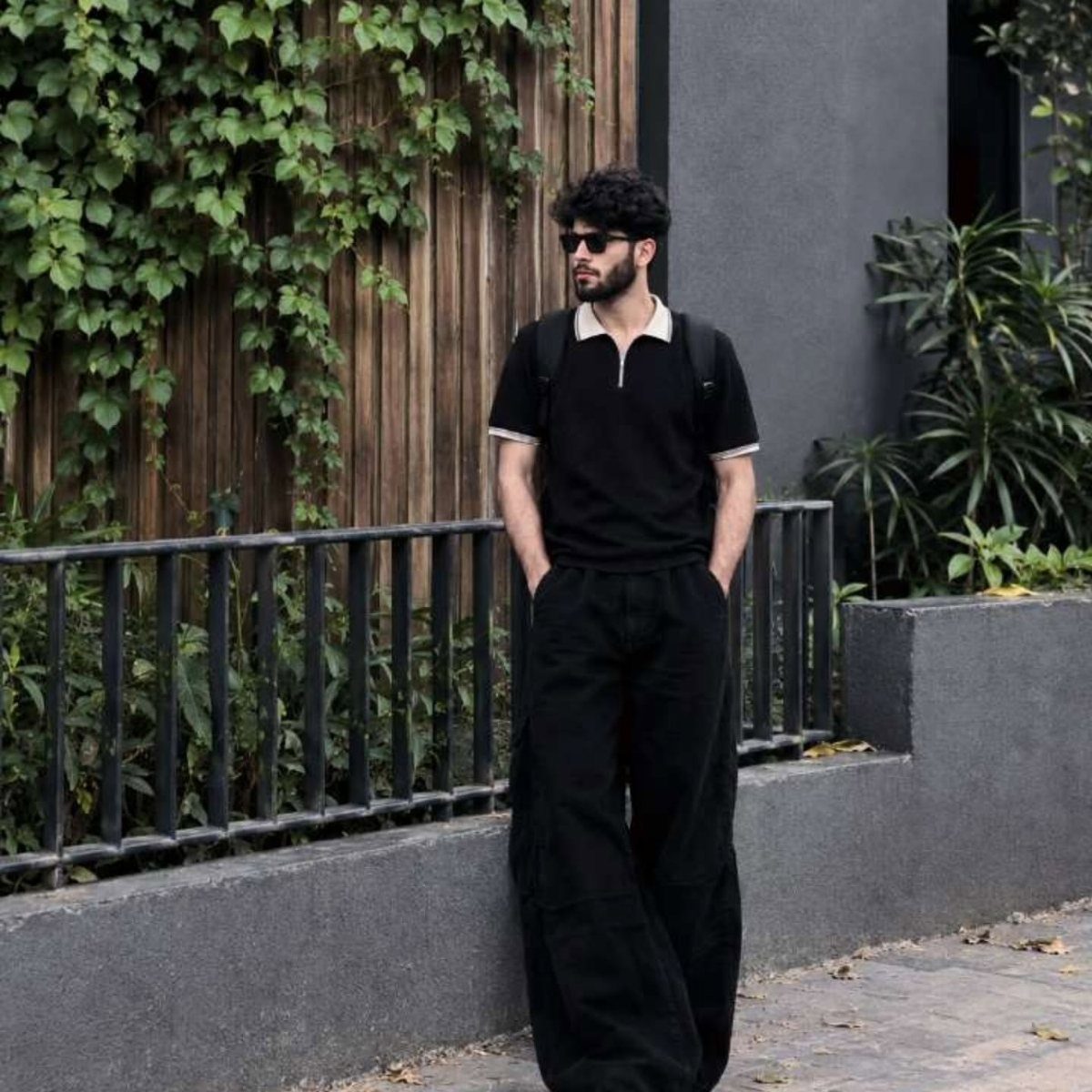 Man wearing a black polo shirt and wide-leg black cargo pants standing by an ivy wall.