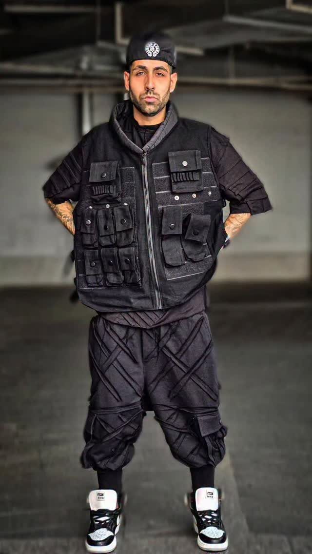 Man in black streetwear outfit with utility vest, patterned cargo pants, and high-top sneakers.
