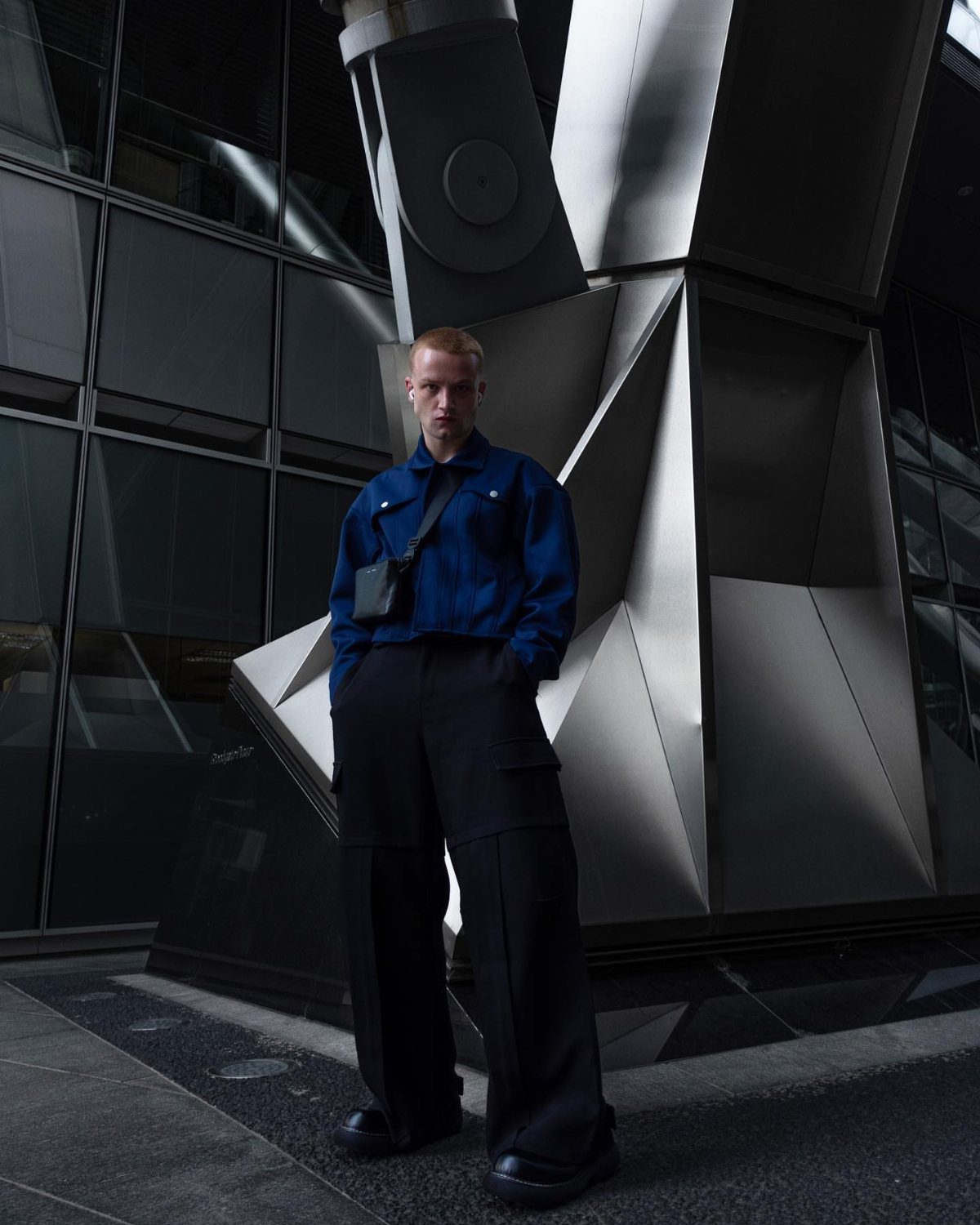 Man wearing a blue cropped jacket and black cargo pants standing by modern architecture.