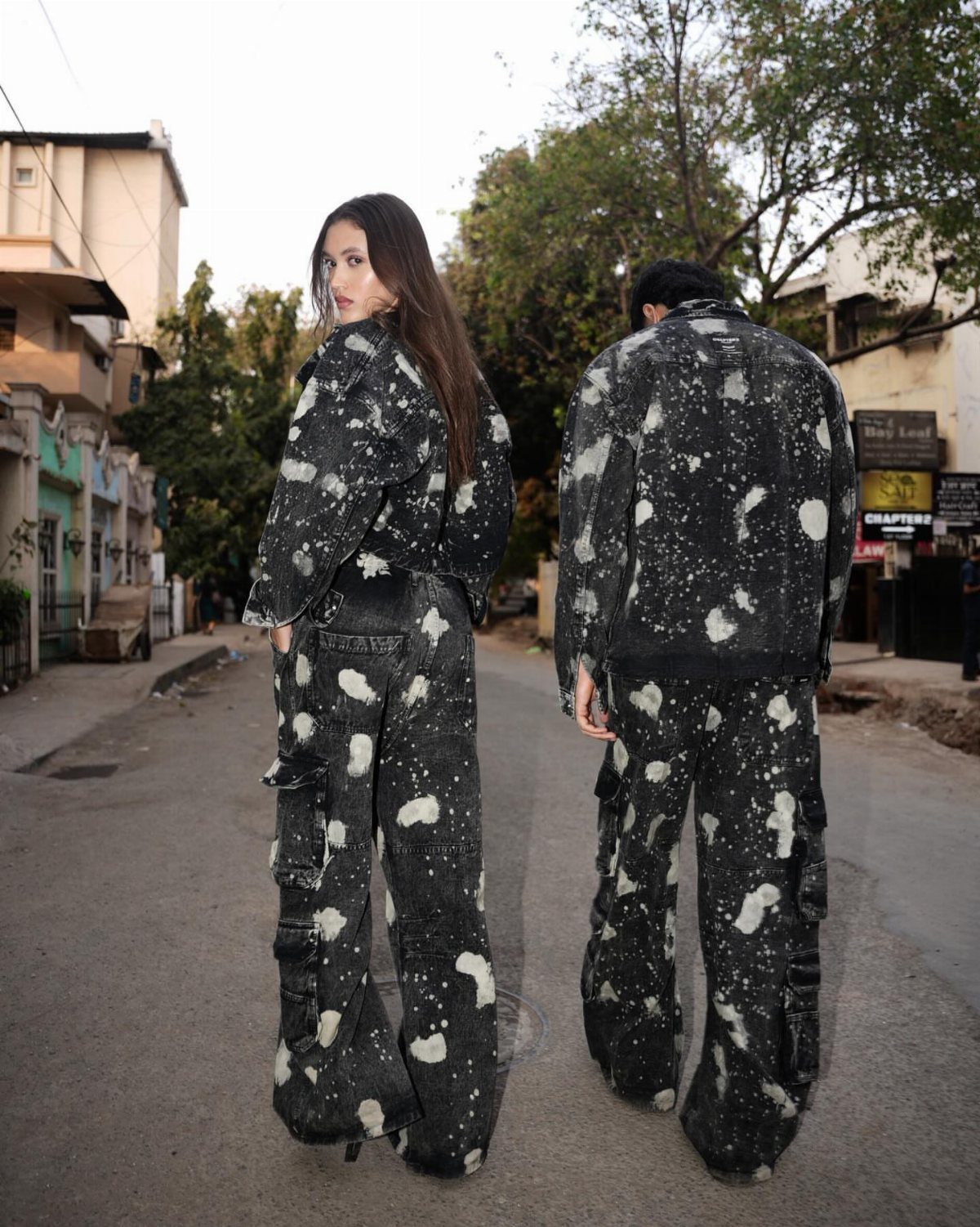 Two people wearing matching black cargo pants and jackets with white paint splatter patterns on a street.
