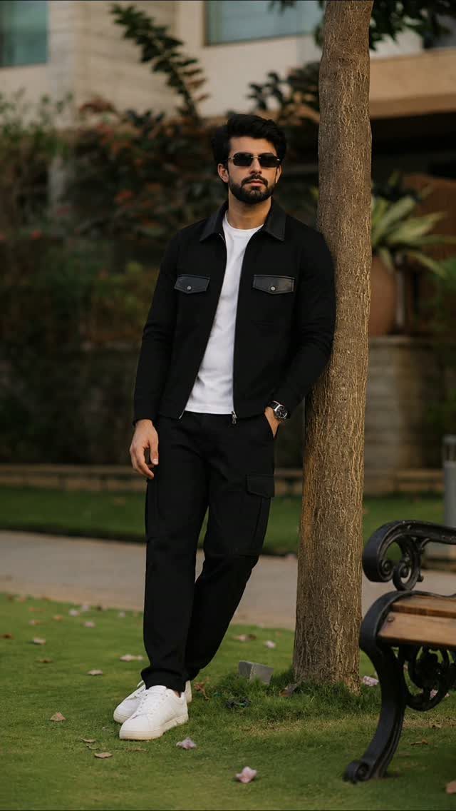 Man in a black utility jacket, white t-shirt, and black cargo pants leaning against a tree.