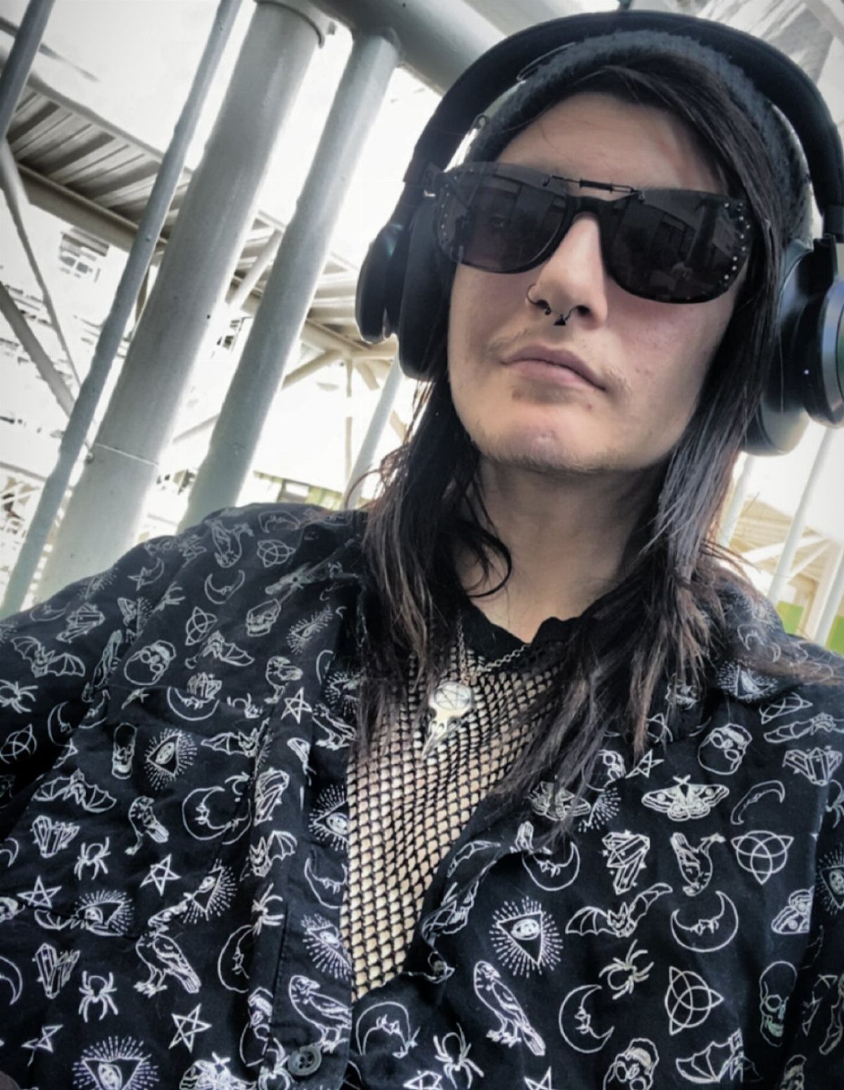 Person with long dark hair wearing large black sunglasses and headphones outdoors.