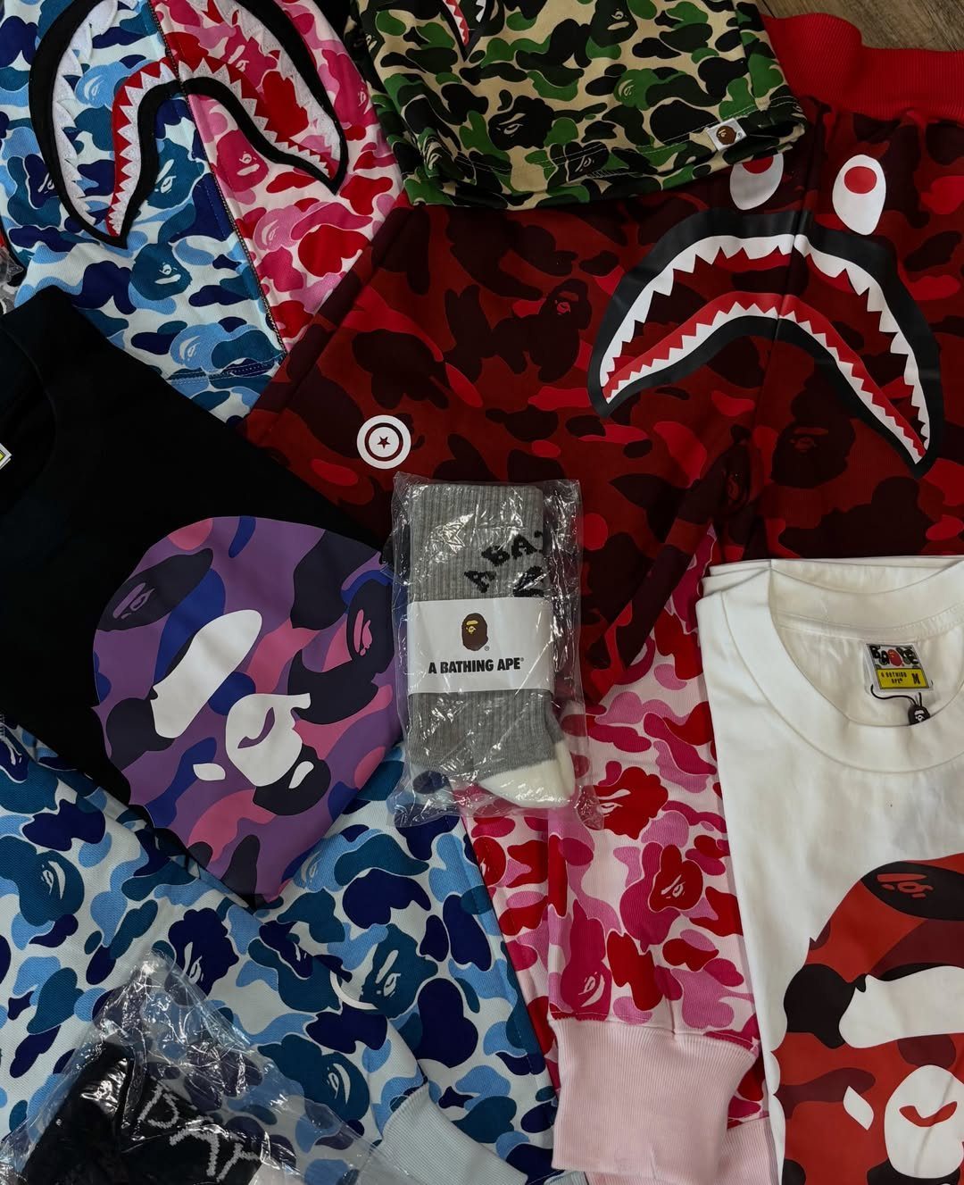 Various BAPE clothing items with colorful camouflage patterns and shark face graphics scattered together.