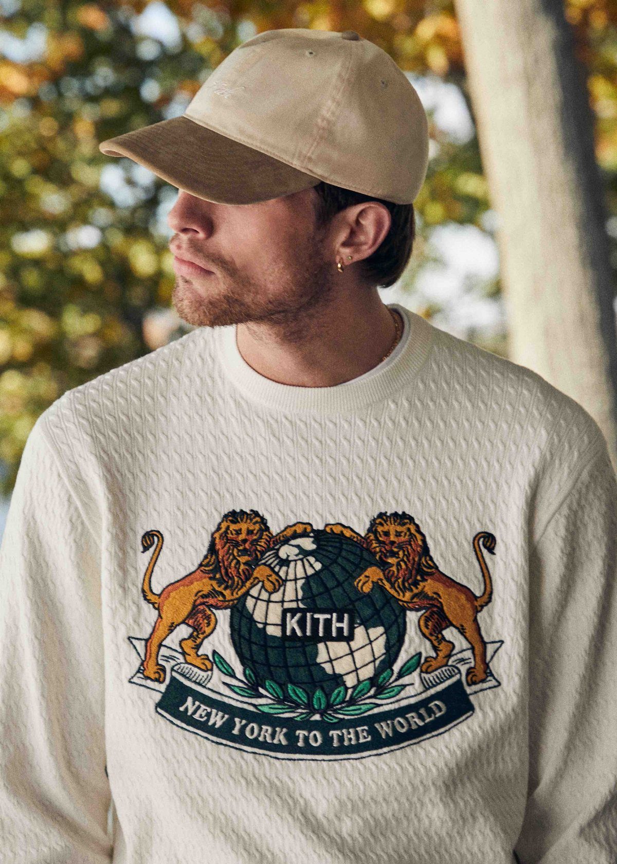 Man wearing a cream KITH sweatshirt with a lion and globe graphic outdoors.