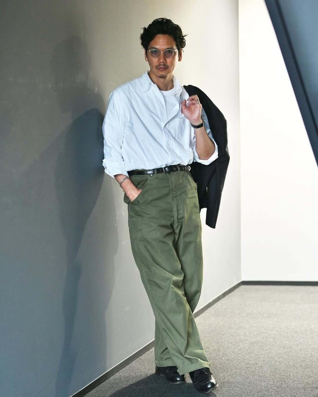 A man in a white shirt and olive green cargo pants leaning against a wall.