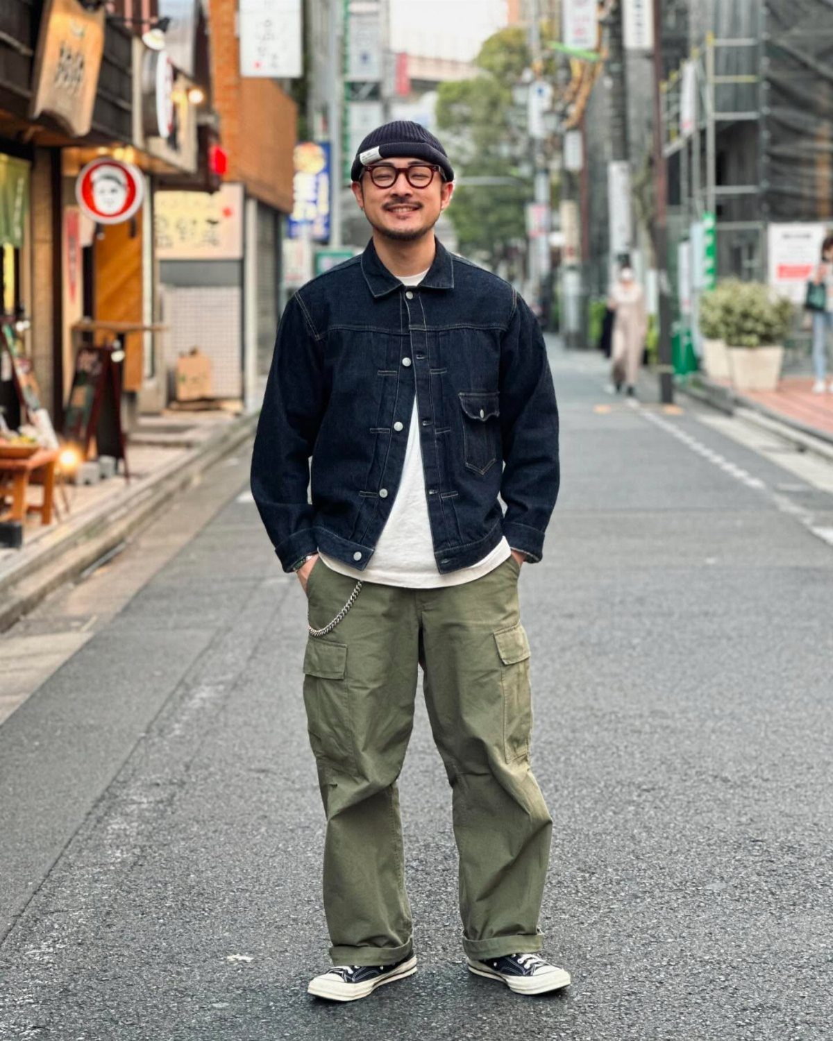 Man wearing a dark denim jacket and olive green cargo pants standing on a city street.