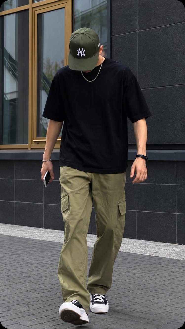 A person wearing a black t-shirt, olive green cargo pants, and a green baseball cap walking outdoors.