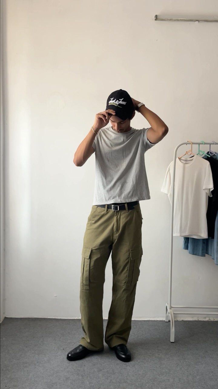 Man wearing olive green cargo pants, a grey t-shirt, and a black baseball cap standing against a white wall.