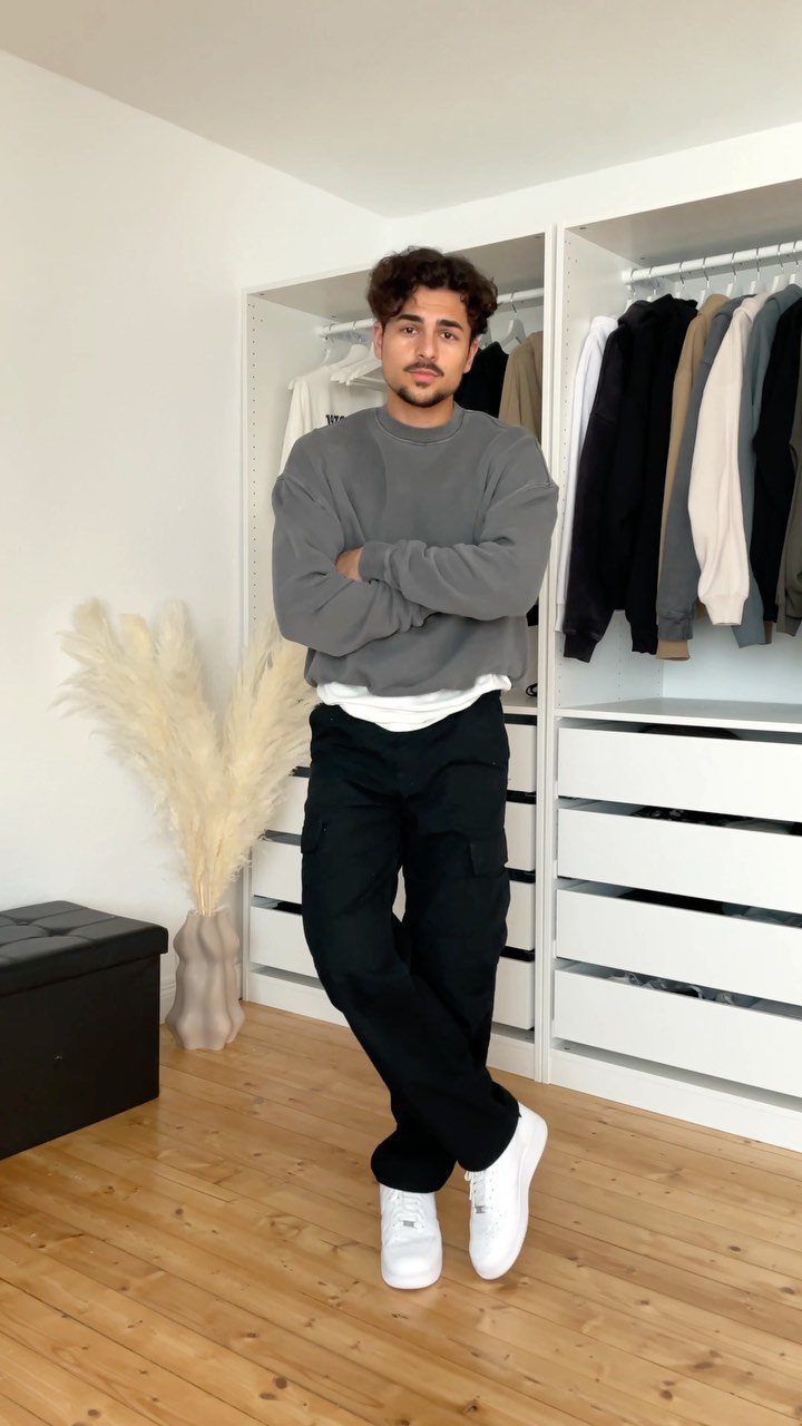 Man posing in a grey sweatshirt, black cargo pants, and white sneakers indoors.