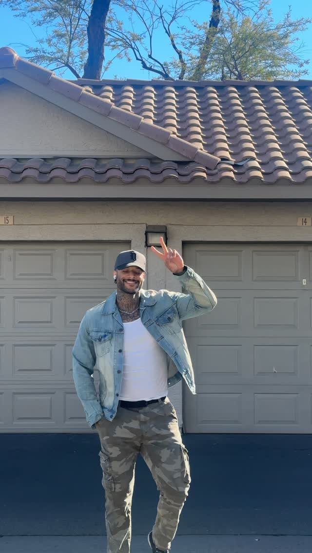 A man smiling in a denim jacket, white tank top, and camo cargo pants.