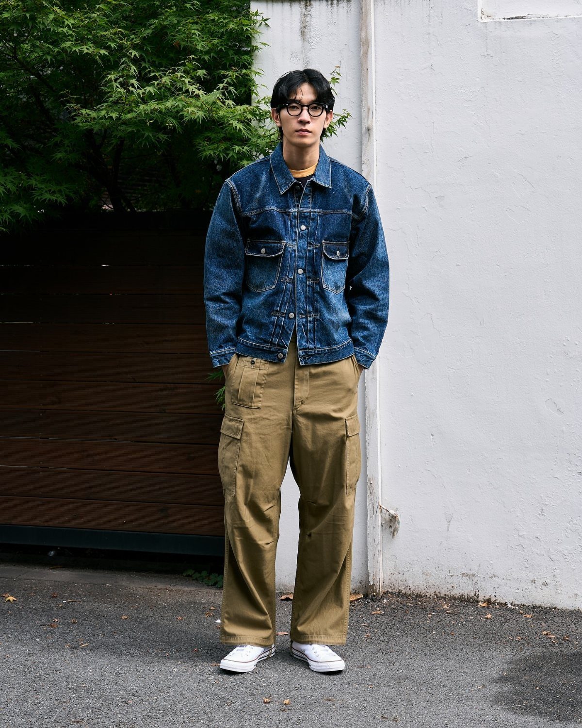 Man wearing a blue denim jacket, tan cargo pants, and white sneakers standing outdoors.