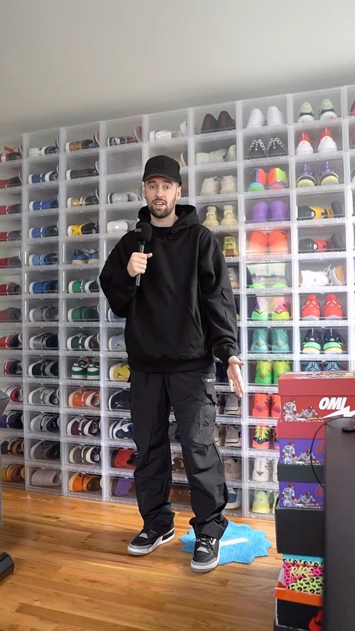 Man in a black hoodie and black cargo pants standing in a room filled with shoes.