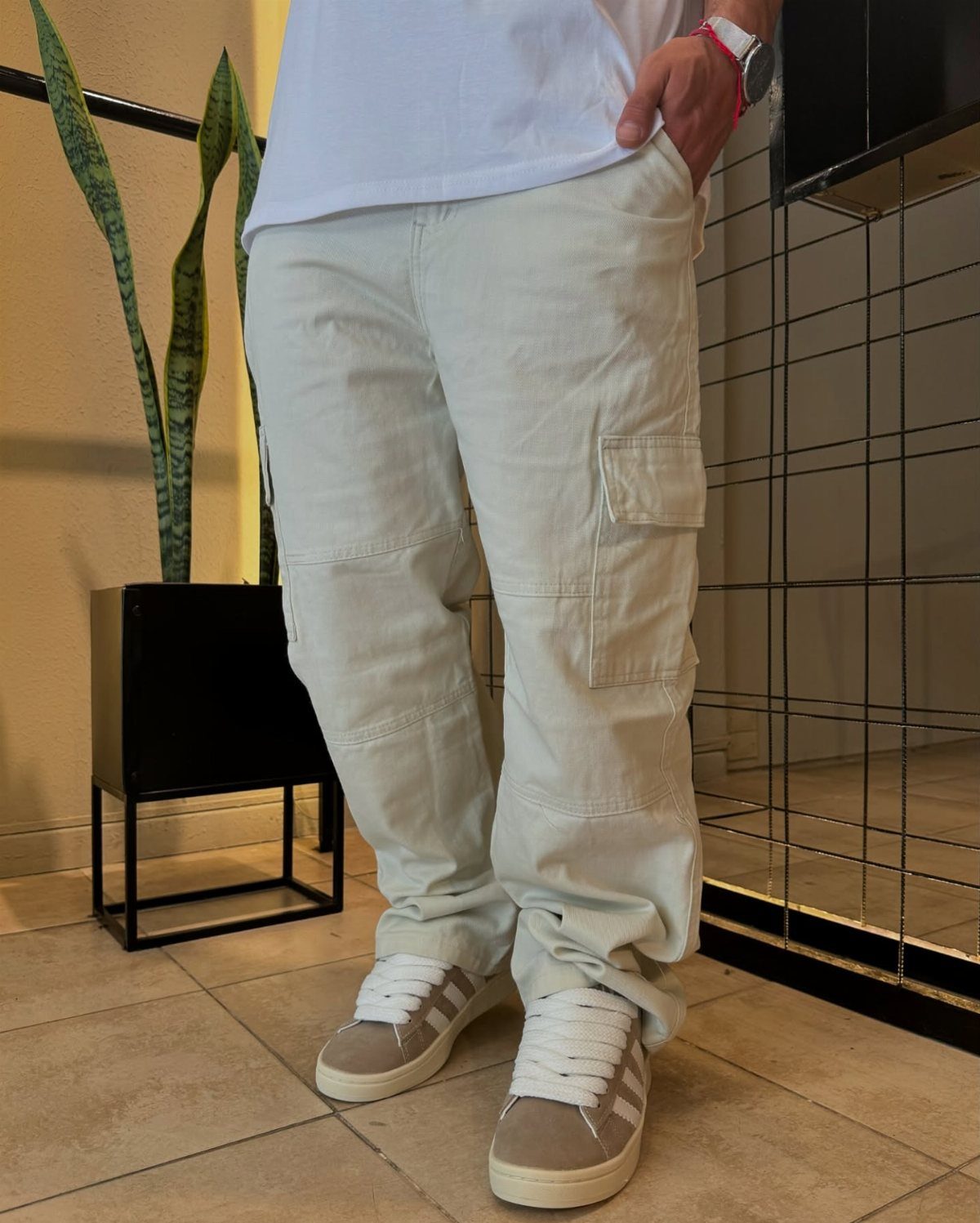 Man standing in a white t-shirt, tan cargo pants, and grey sneakers near a potted plant.