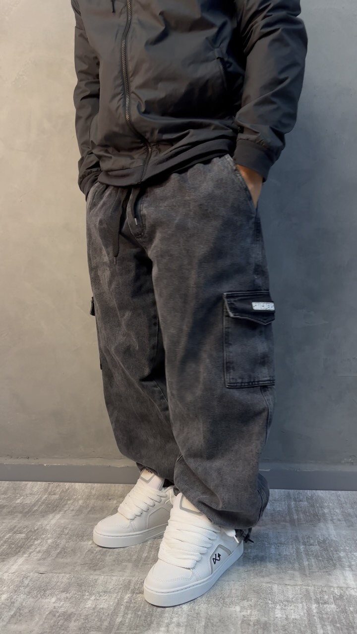 Person wearing dark grey washed cargo pants and white sneakers against a grey wall.