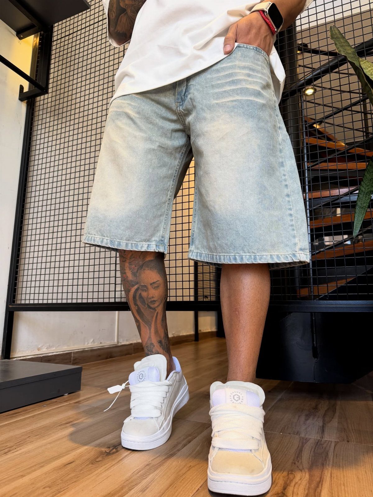 Person wearing light blue denim Bermuda shorts and white sneakers standing on a wooden floor.