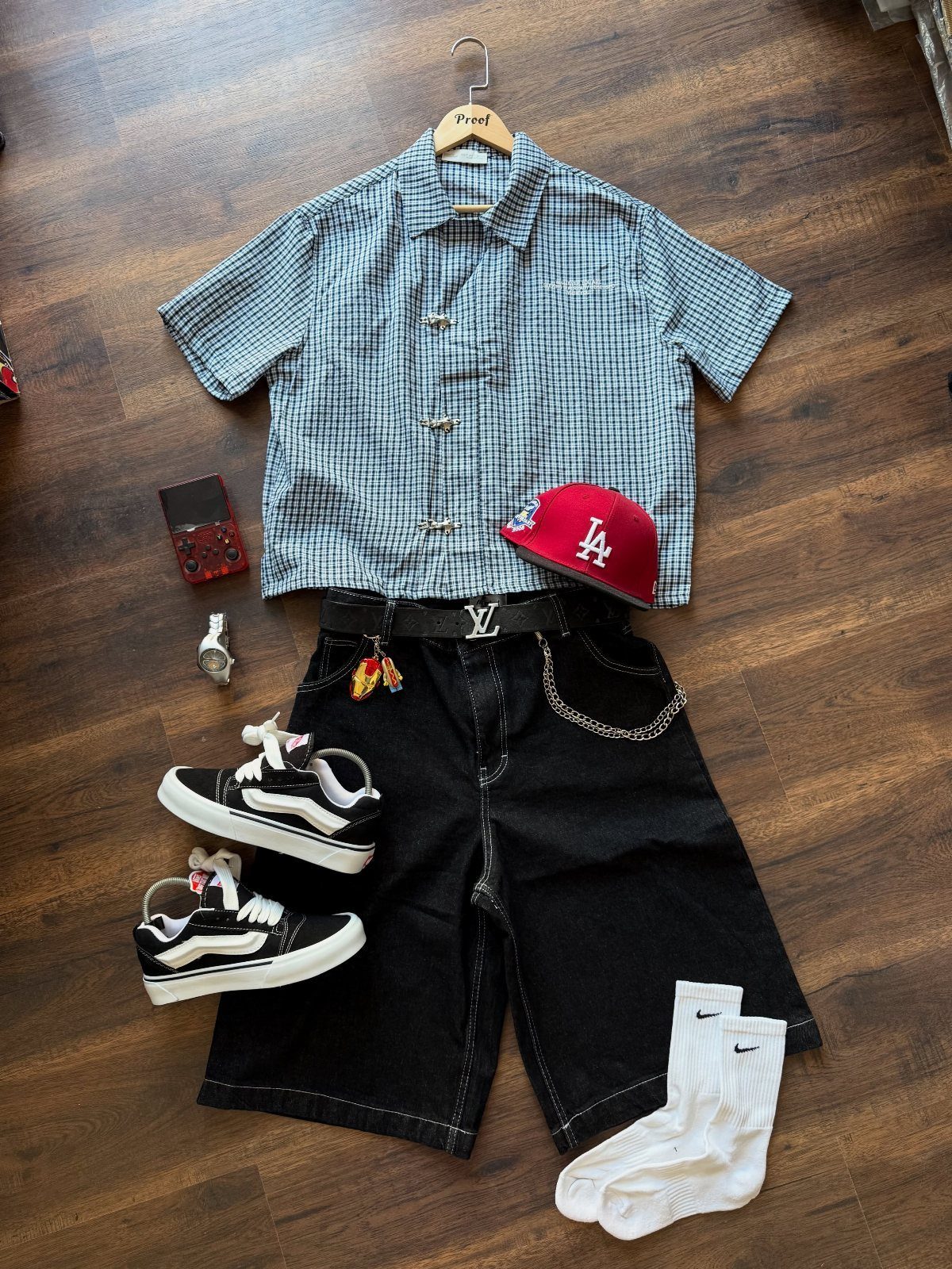 Flat lay of casual streetwear including a checkered shirt, black shorts, red cap, and designer belt.