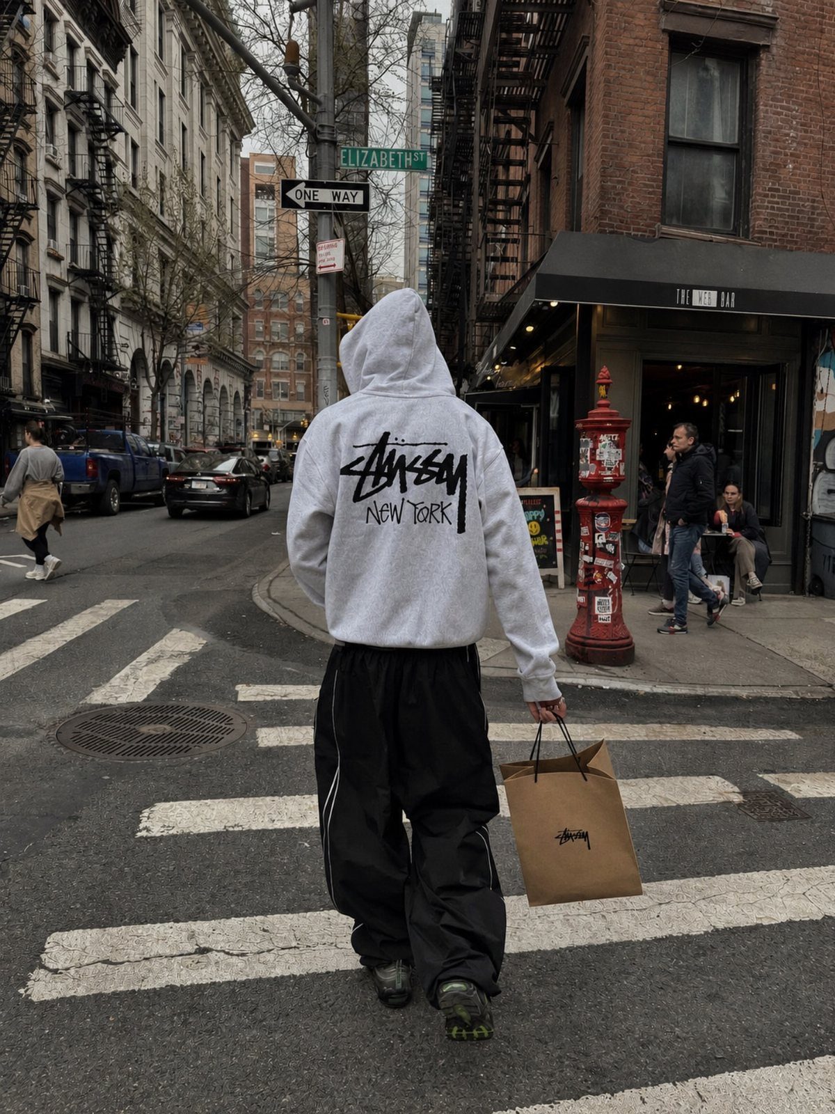 Person in a grey graphic hoodie and black pants walking across a city street crosswalk.