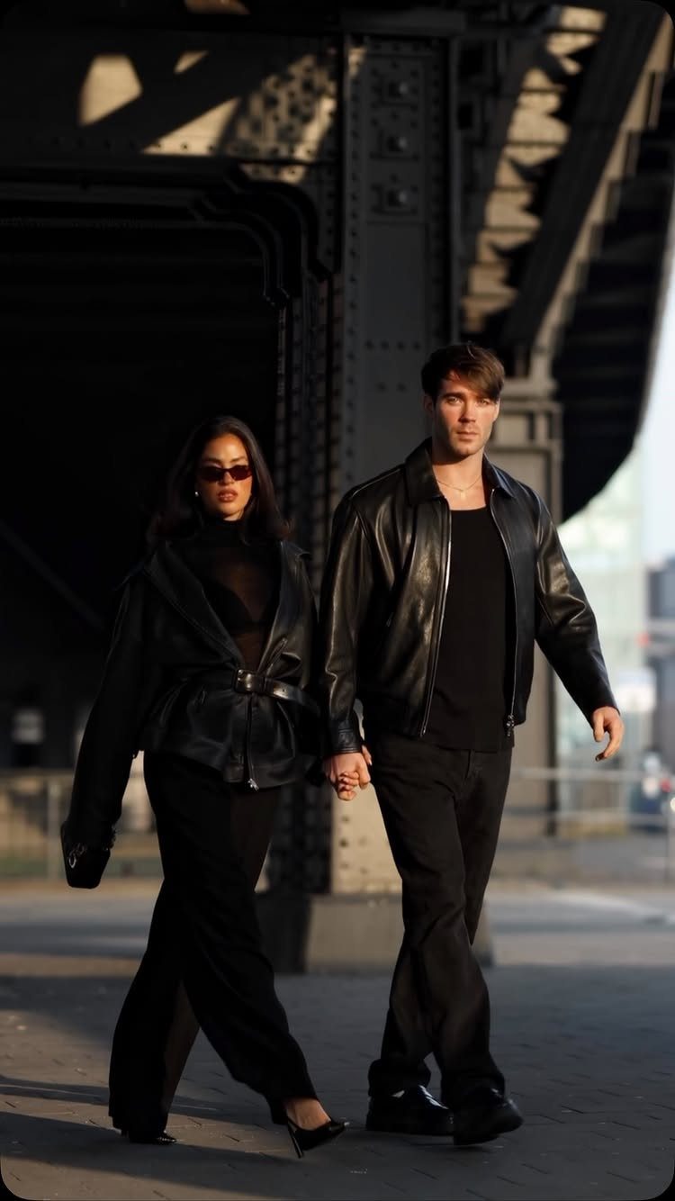 A man and woman walking in black leather jackets and dark trousers under a bridge.